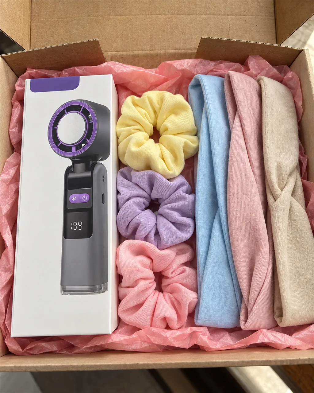 El ventilador de mano en su caja junto a scrunchies y diademas de tela de varios colores: son los colores para elegir; el regalo es un scrunchie y una diadema